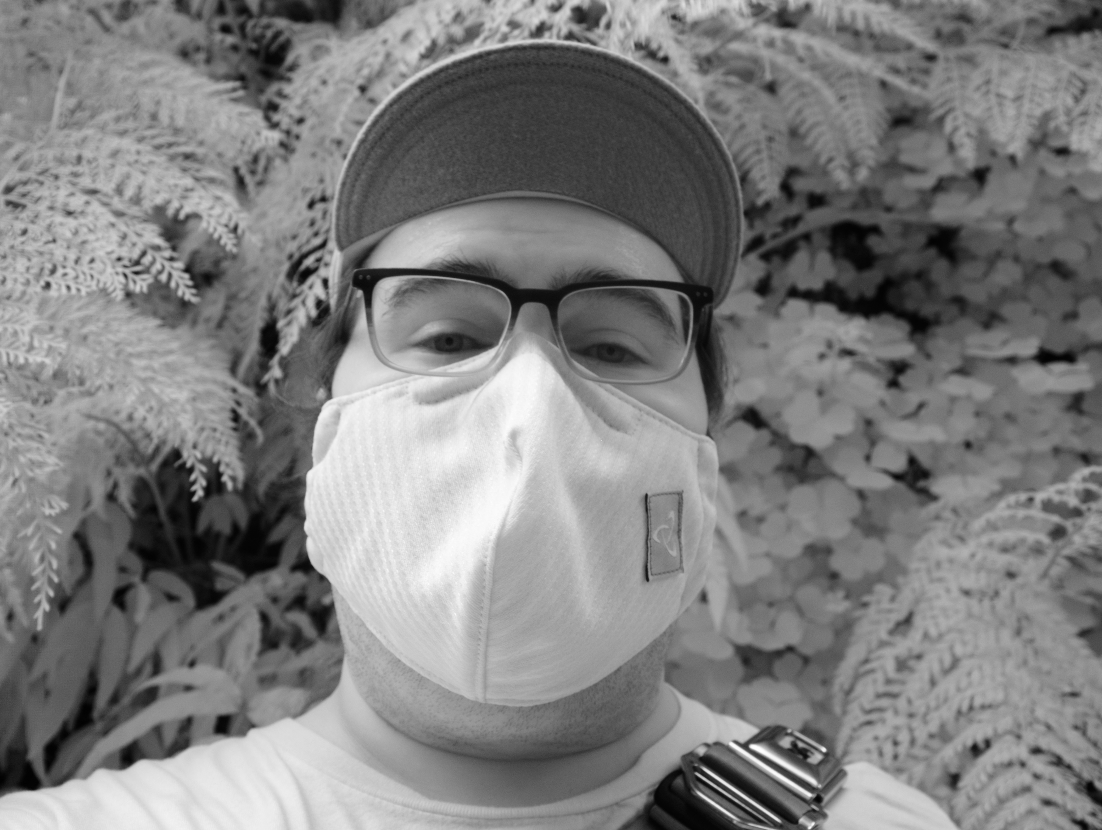 Black and white infrared photo of a nerd wearing a mask, glasses, brimmed cap, and a chrome bag, standing in front of a "green wall" plant installation